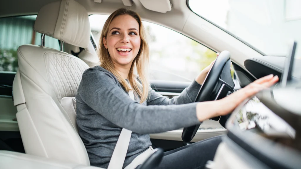 A smiling woman in a gray sweater sits in the driver's seat of a luxury car, looking out the side window.