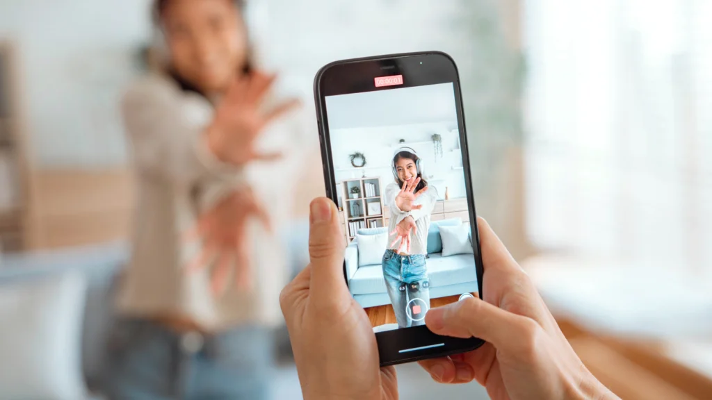 A woman filming herself on a smartphone in a bright room, wearing headphones. She gestures playfully with a smile.