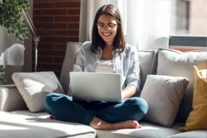 A smiling woman with glasses sits cross-legged on a sofa, using a laptop to find side hustles for women. She’s surrounded by cushions.