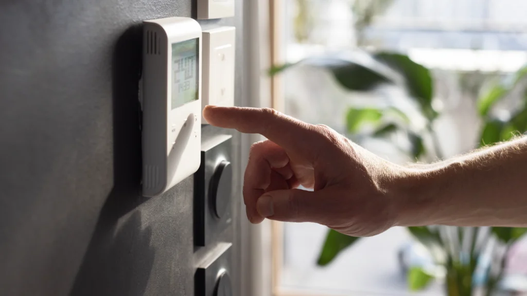 A hand adjusting a digital thermostat mounted on a dark wall, showing temperature settings.
