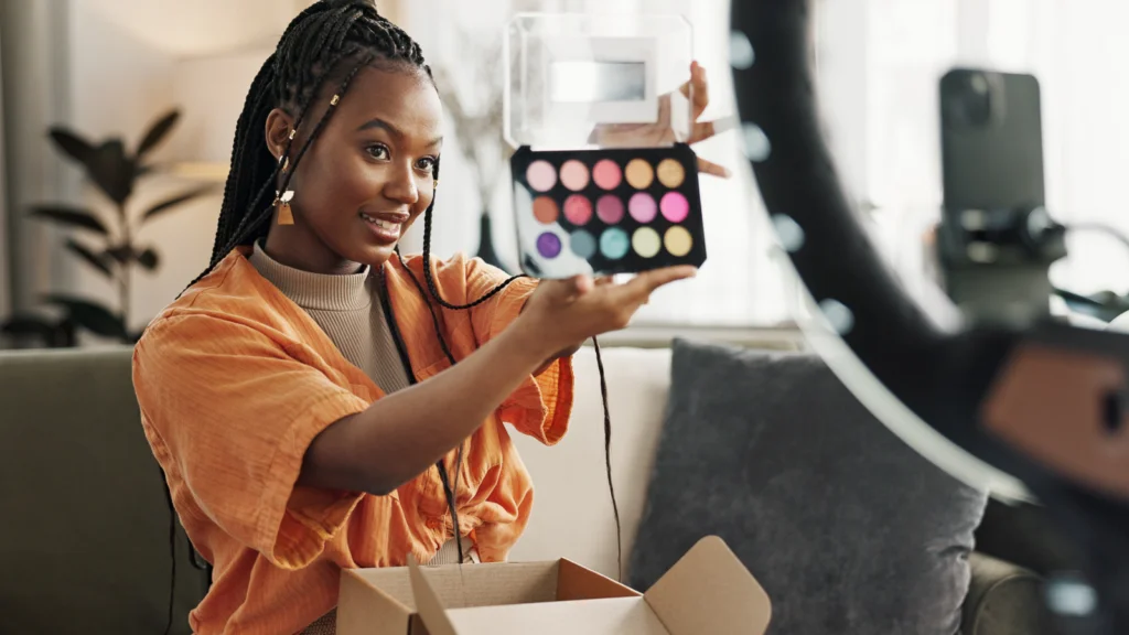A smiling woman in an orange shirt presents a colorful eyeshadow palette to a camera. She sits on a sofa.