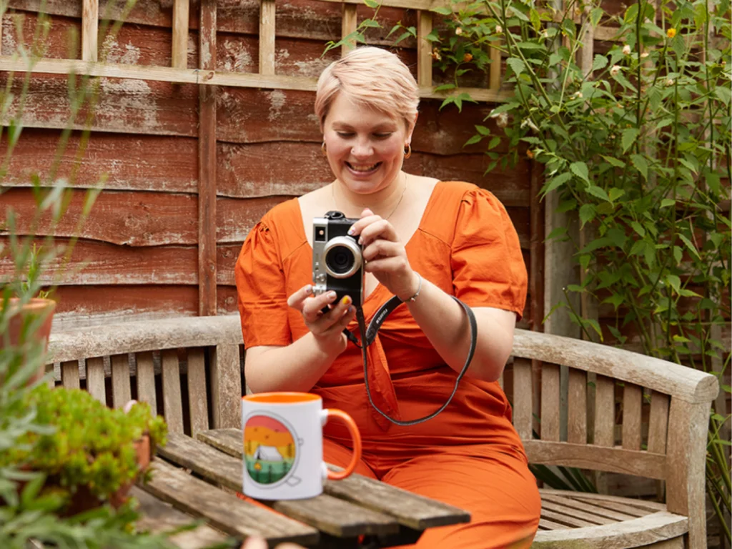 A woman in an orange dress smiles as she takes a picture of a mug with a digital camera, seated on a wooden bench in a garden.