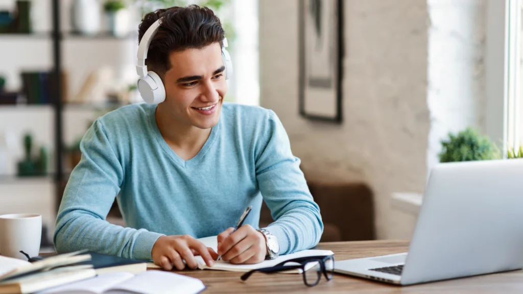 A man in a blue sweater, wearing headphones, smiles while writing in a notebook at a wooden table. An open laptop and books are nearby.