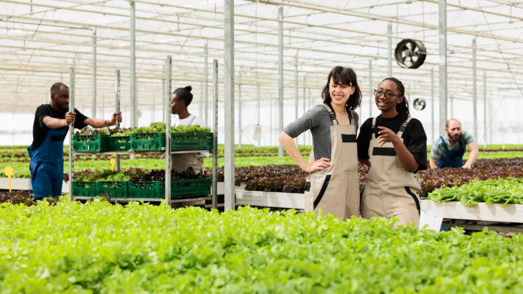 Two women in aprons smile in a greenhouse filled with green plants. Three people work in the background.