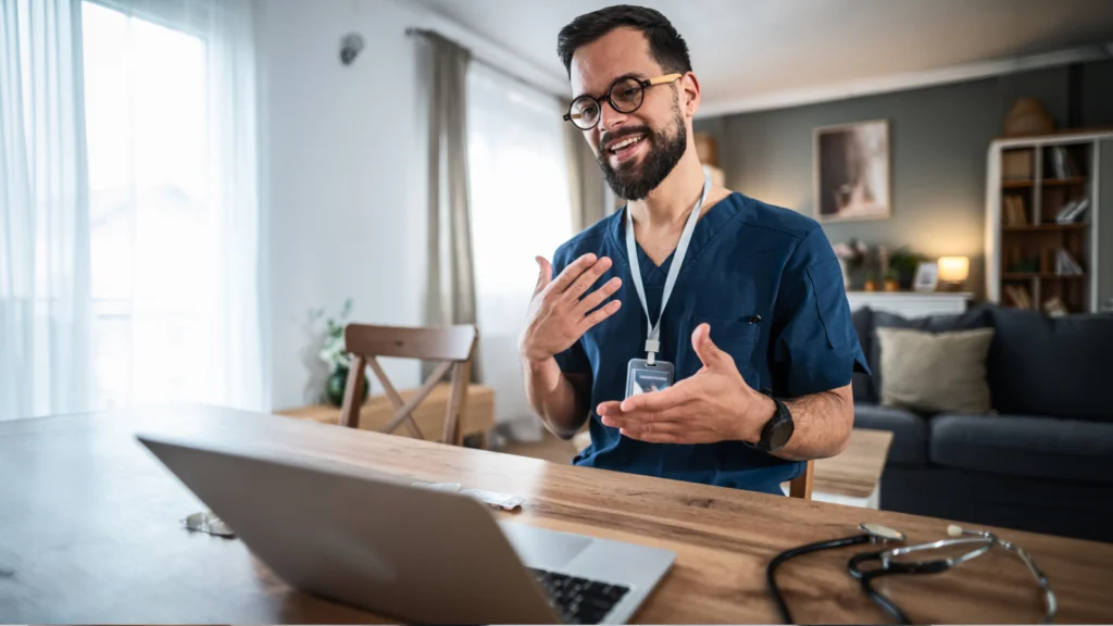 A smiling male doctor with glasses and a beard conducts a telehealth appointment on a laptop at home.