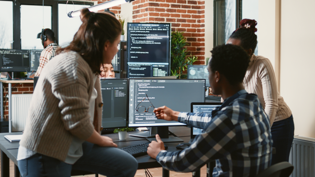 Three people collaborate in a tech office, discussing code on multiple computer screens.
