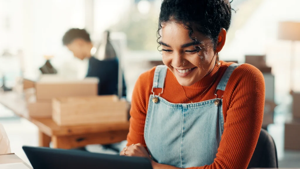 A woman with curly hair smiles while looking at a laptop. She wears a red sweater and denim overalls.