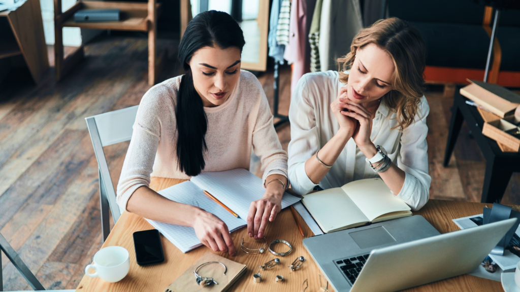 Two women are sitting at a wooden table, discussing jewelry designs. Open notebooks, jewelry pieces, a laptop, and a cup are visible.