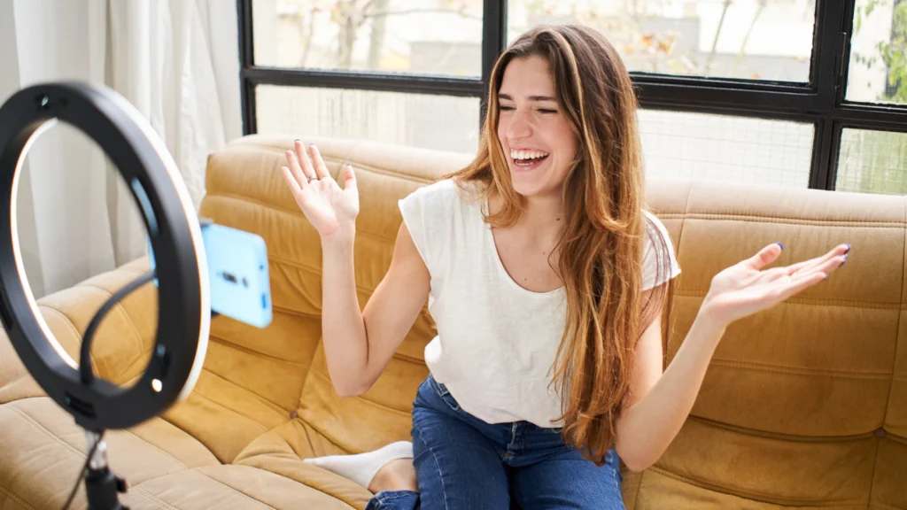 A woman sits on a sofa, smiling and gesturing in front of a phone mounted on a ring light.