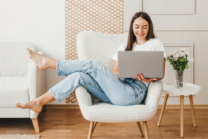A smiling woman in a white t-shirt and blue jeans is sitting on a chair with a laptop, searching for how to start an Etsy shop.