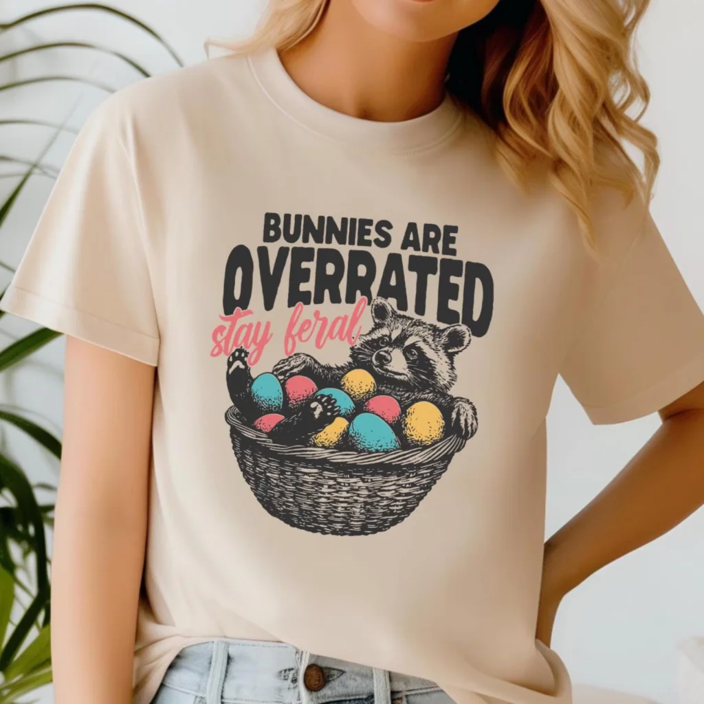 A woman wearing a beige shirt featuring a raccoon in a basket of colorful Easter eggs. Text reads "Bunnies are overrated, stay feral".