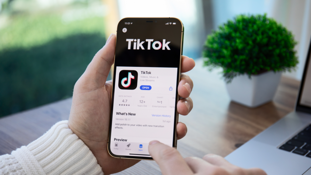 Hand holding a phone displaying the TikTok app in the app store. A blurred green plant and a laptop in the background.