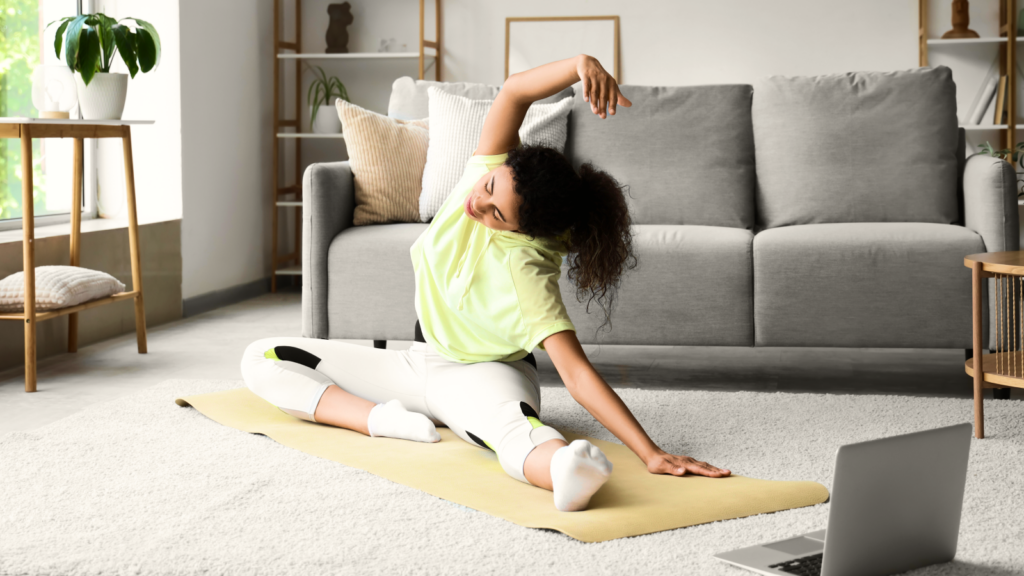 A woman in workout clothes stretches on a yoga mat in a cozy living room, with a laptop nearby.