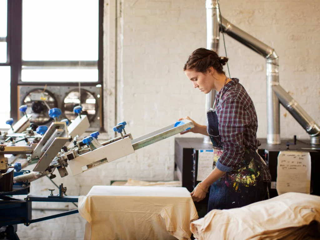 01 What is the Printify order fulfillment process A woman in a plaid shirt and paint-splattered apron operates a printing press in a workshop with exposed pipes.