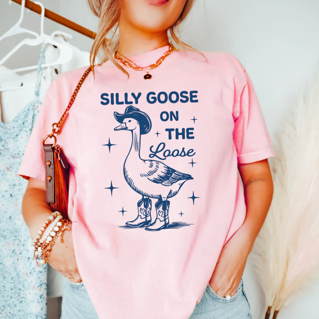 A woman in a pink t-shirt with a cartoon of a goose wearing a cowboy hat and boots. The text reads "Silly Goose on the Loose".