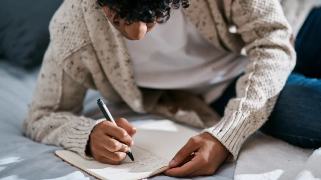 A man with curly hair is lying on a bed, wearing a cozy sweater, focused on writing in a notebook.