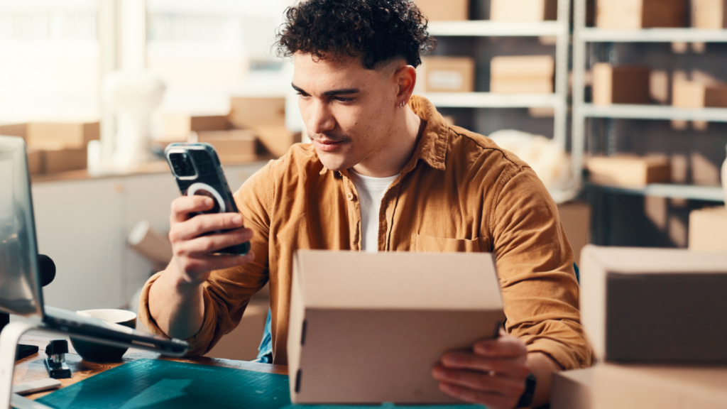 A man in a brown shirt holds a cardboard box while looking at his phone, surrounded by packed boxes on shelves.