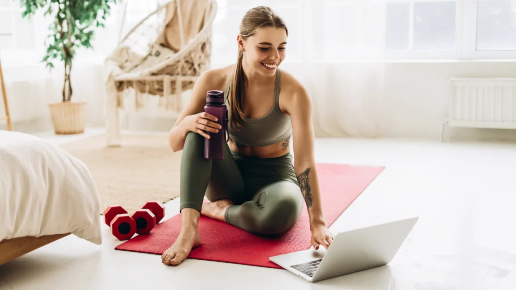 A woman in workout attire sits on a yoga mat, holding a purple water bottle, smiling at a laptop. Red dumbbells lie nearby.