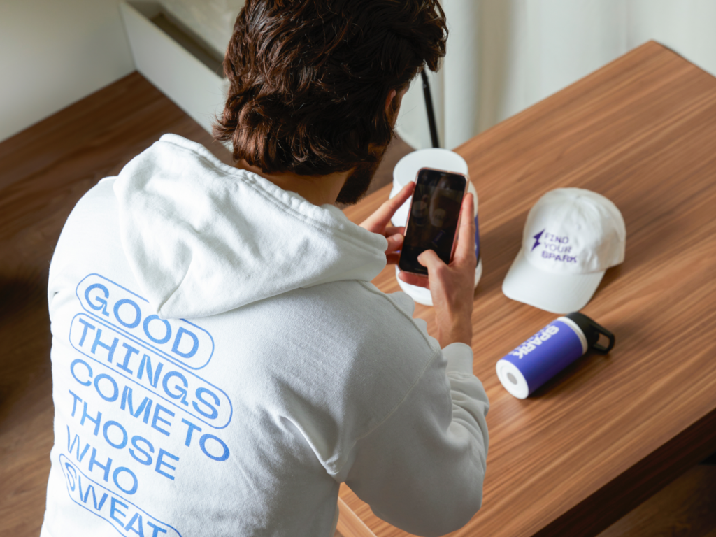 A man in a white “Good things come to those who sweat” hoodie. He takes a photo with a smartphone beside a cap and a water bottle.