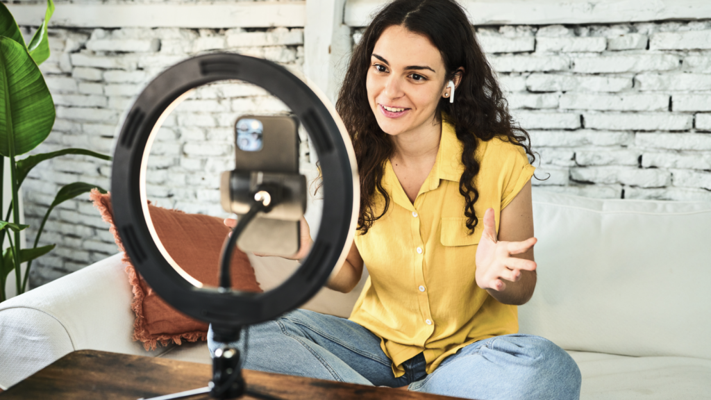 A woman in a yellow shirt sits on a couch, smiling and gesturing as she records with a smartphone on a ring light stand. Brick wall background.