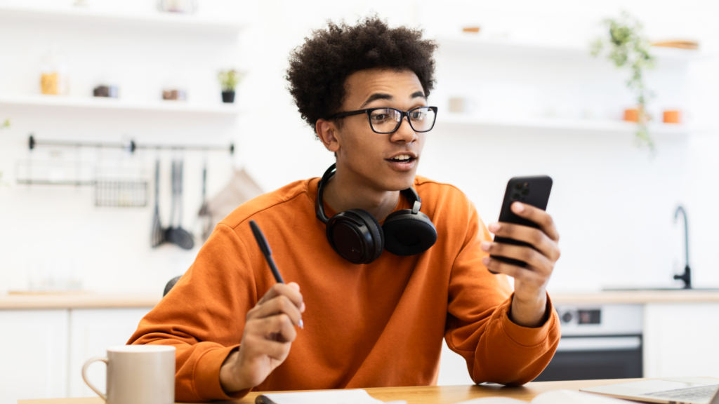A young man with glasses and headphones, holding a smartphone in one hand and a pen in the other, searching “Is TikTok shop worth it”.