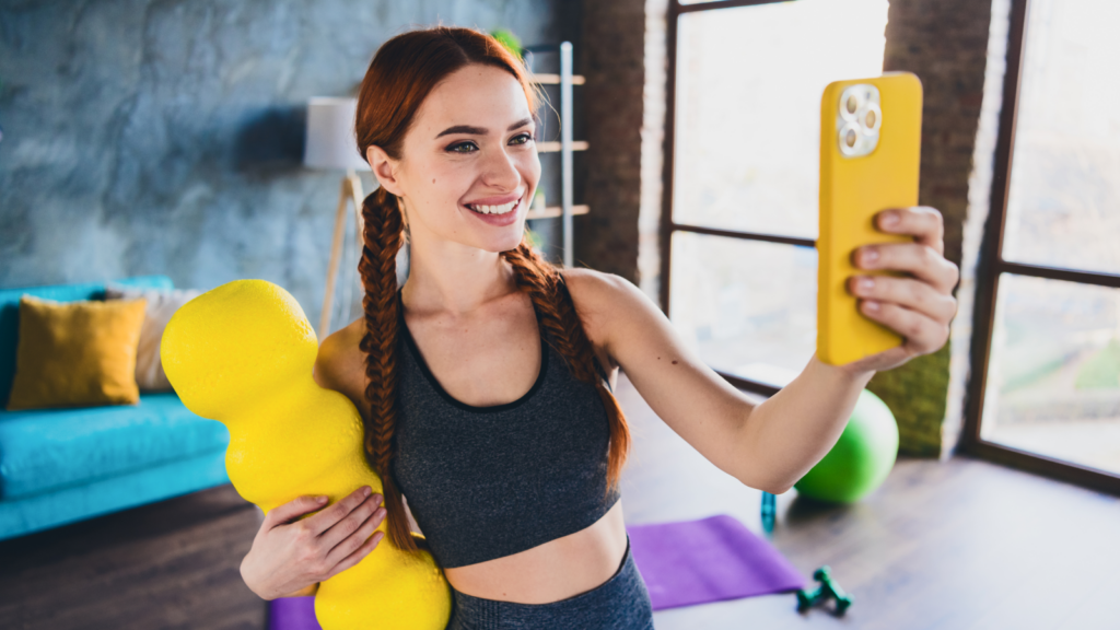 A young woman with red braided hair smiles while taking a selfie in a modern living room. She holds a yellow foam roller.