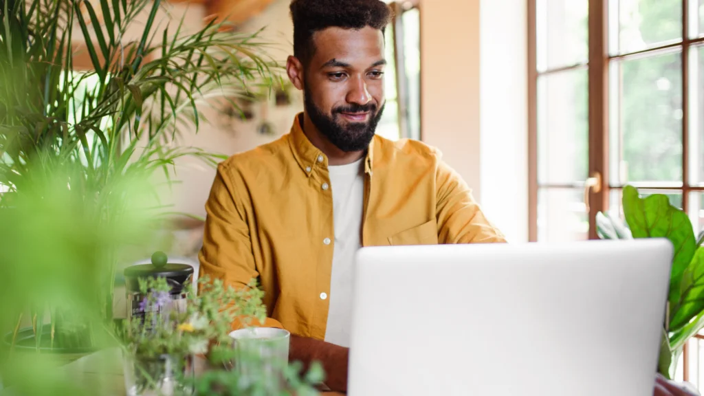 A man in a yellow shirt works on a laptop surrounded by lush green plants, near a sunlit window.