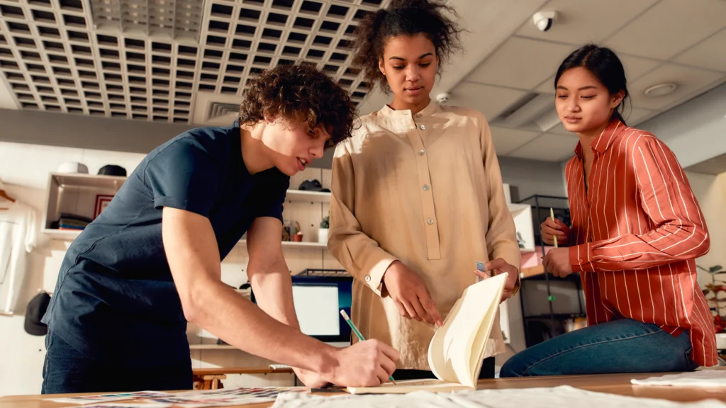 04 What are the stages of custom product production Three young adults collaborate in a modern office. One writes on a notebook, while the others observe and discuss.