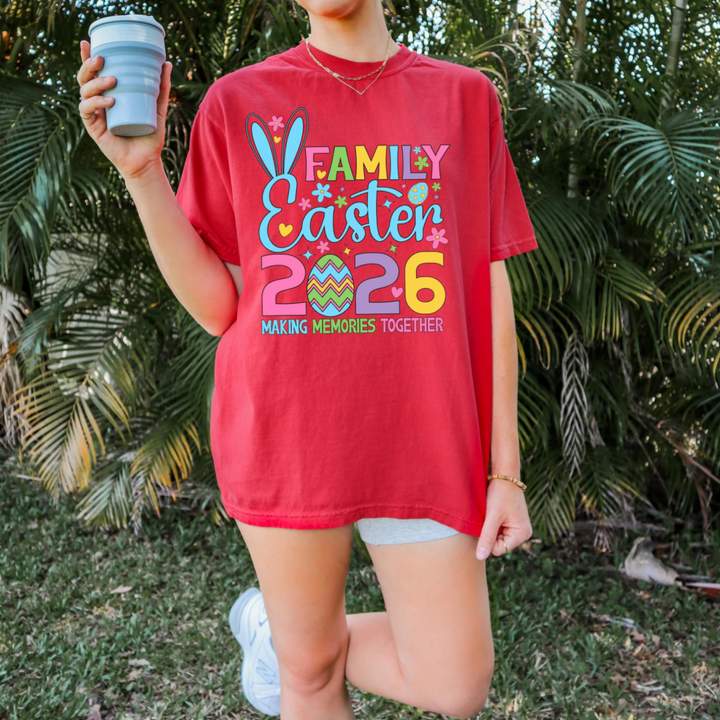 A person in a red t-shirt reading "Family Easter 2026, Making Memories Together," with colorful text and bunny ears, stands in a garden with a drink.