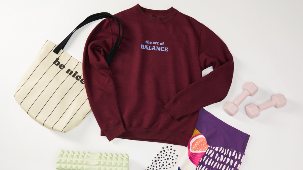 A flat lay image featuring a burgundy sweatshirt with the text "the art of BALANCE," a striped tote bag, pink dumbbells, and colorful leggings.