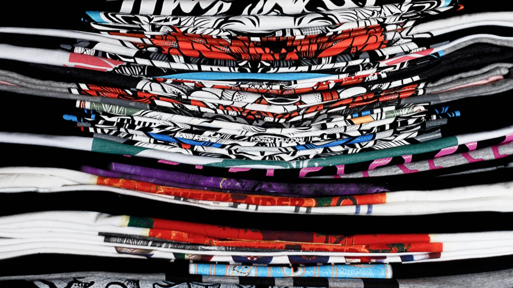 05 What decoration methods does Printify use A stack of colorful, patterned t-shirts in various designs and hues, neatly folded.