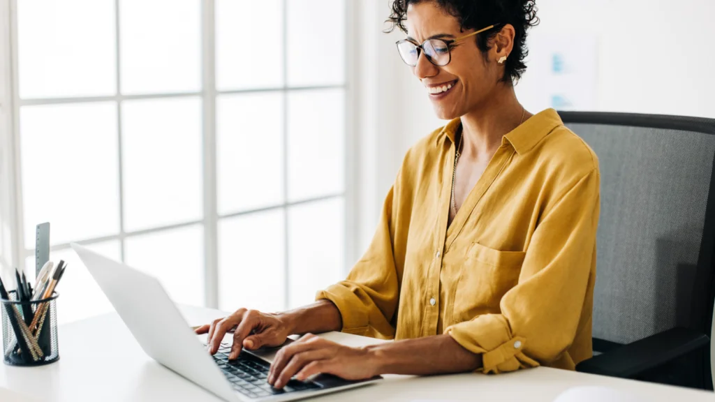 A woman in a yellow shirt smiles as she types on a laptop at a bright desk, with soft, natural light from large windows.