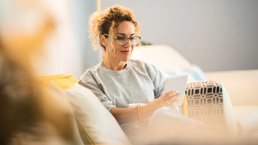 A woman with curly hair and glasses sits on a couch, relaxed, reading a tablet. She wears a gray sweater.
