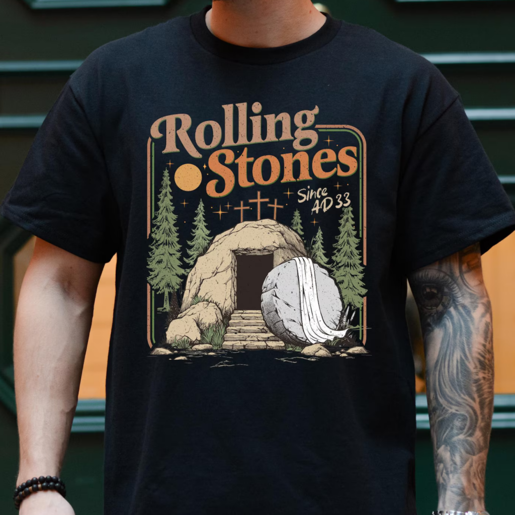 A man wearing a black t-shirt with a design featuring "Rolling Stones," a rock tomb entrance with a rolled stone and trees.