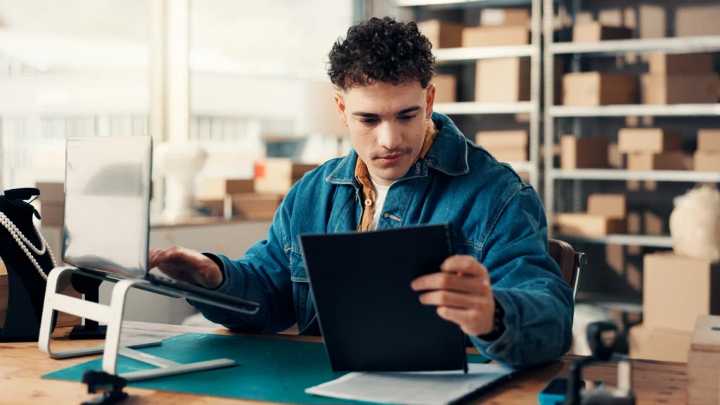 A man in a denim jacket is working at a desk with a tablet and a laptop in a warehouse. Shelves with boxes are visible in the background.