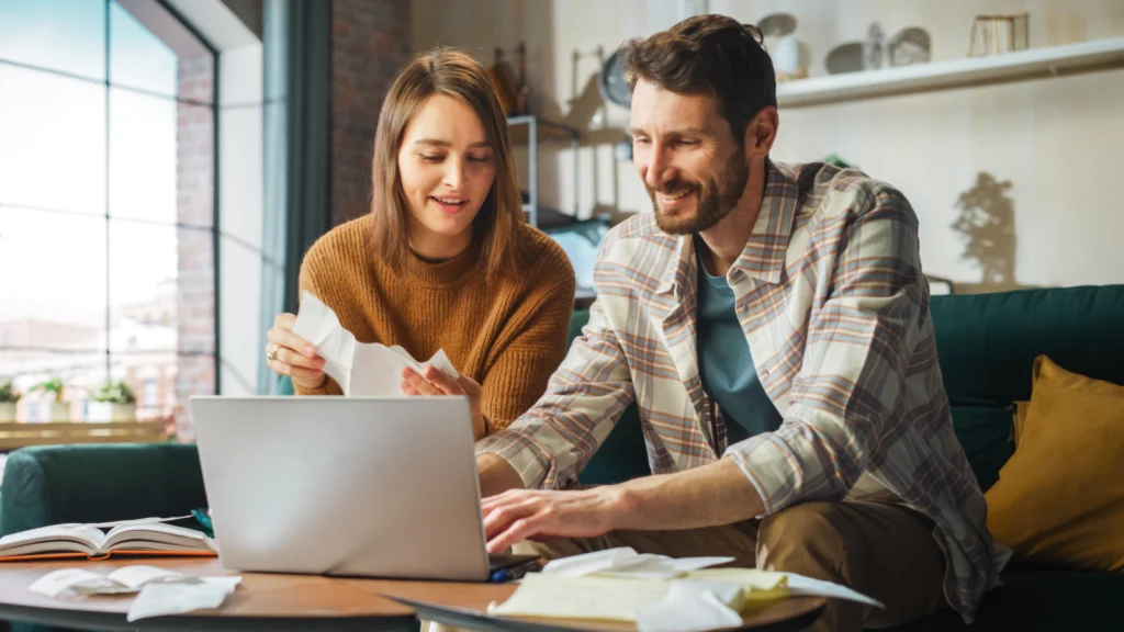 07 What support does Printify offer throughout the order to delivery journey A couple sits at a table, smiling and working together on a laptop. They are surrounded by papers.