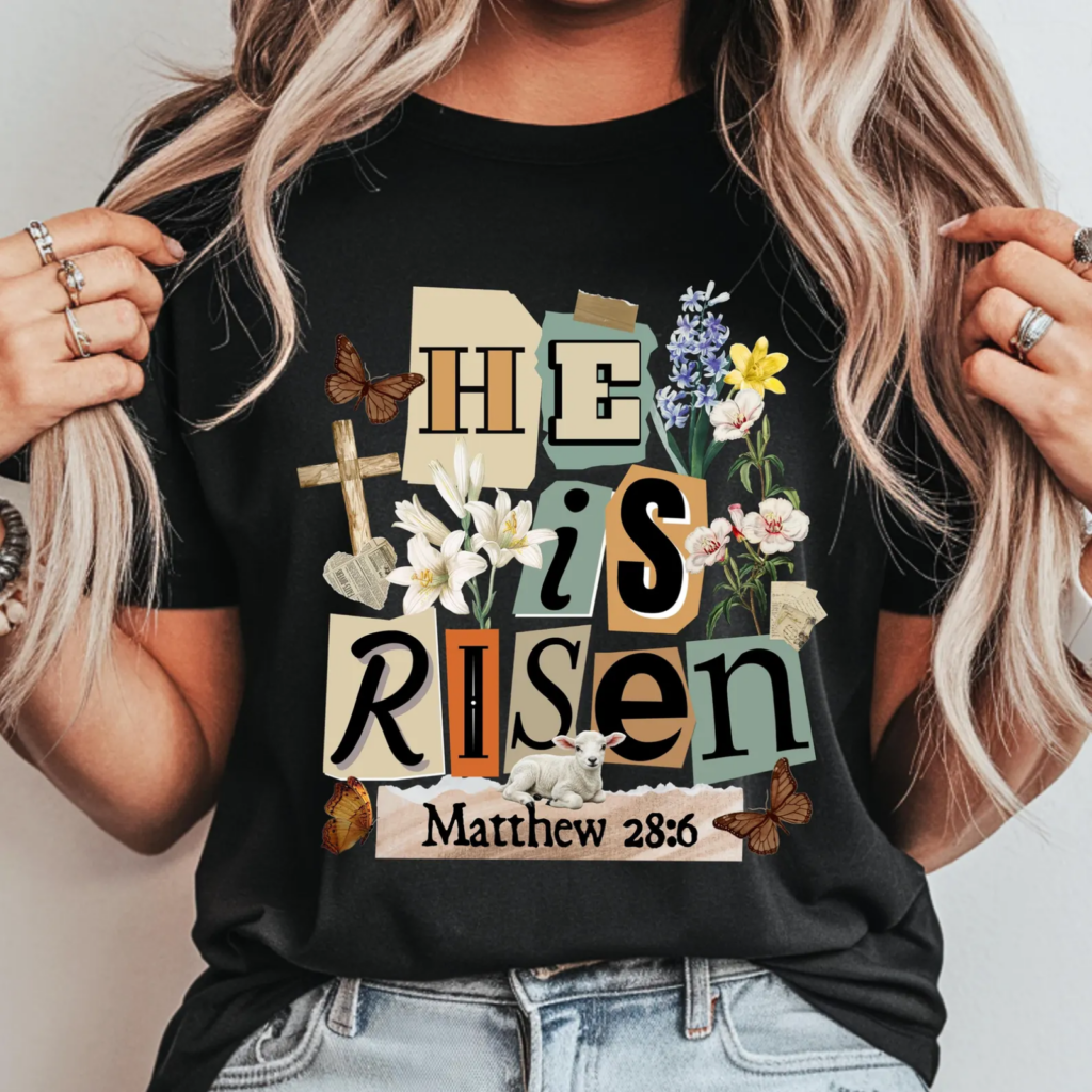 Woman wearing a black shirt with "He is Risen" in colorful letters, flowers, butterflies, a lamb, and a cross, with "Matthew 28:6" at the bottom.