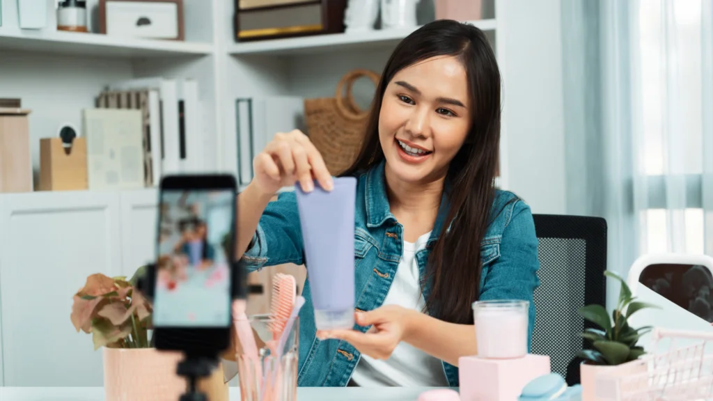 A cheerful woman records a video holding a purple skincare product, surrounded by beauty items on a desk.