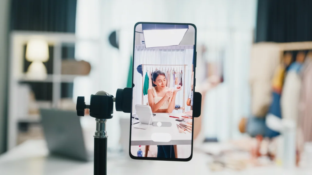 A woman livestreaming a makeup tutorial on her phone in a bright room.