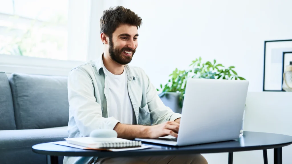 A man smiles while typing on a laptop at a modern living room table.