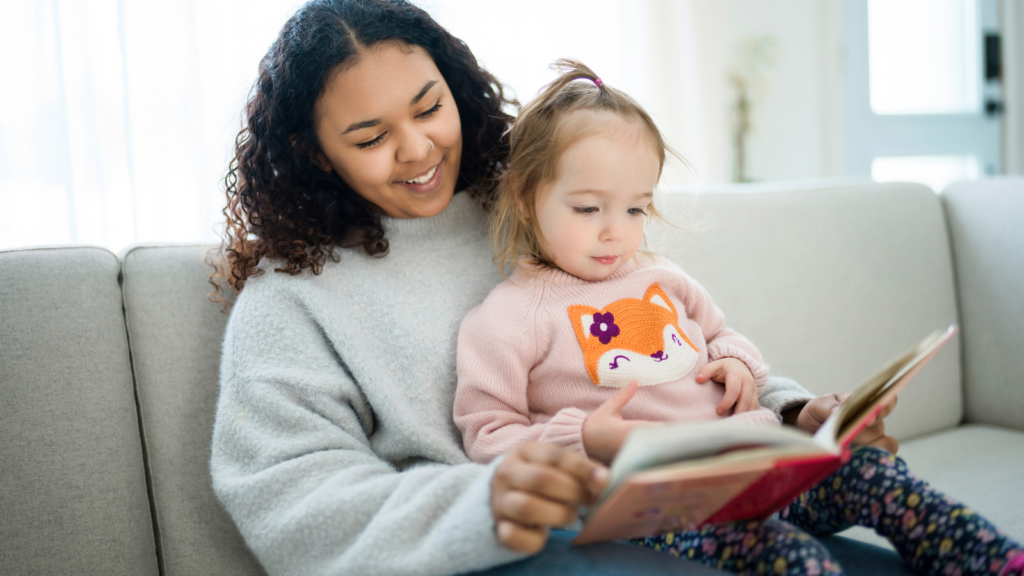 A smiling woman in a gray sweater reads a book with a young girl in a pink fox sweater. They sit on a light gray sofa.