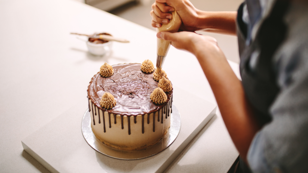 A person decorates a chocolate-drizzled cake with a piping bag, adding swirls on top.