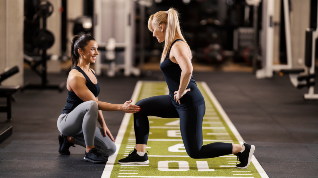 A trainer in a black tank top and gray leggings kneels while instructing a woman in a lunge on gym turf.