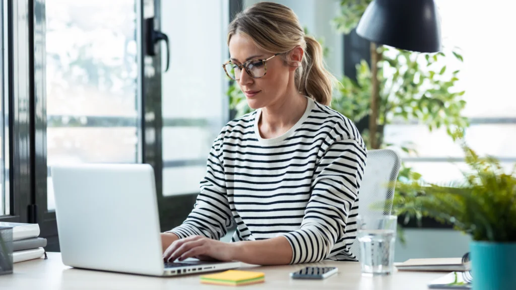 Best ways to make money with AI A focused woman in glasses and a striped shirt works on a laptop at a modern desk with plants and a window view.