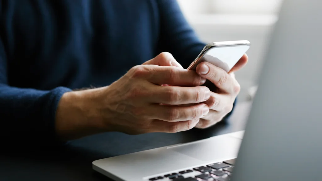 Build AI chatbots for businesses A person in a dark sweater uses a smartphone, focusing intently while sitting at a desk with a laptop.