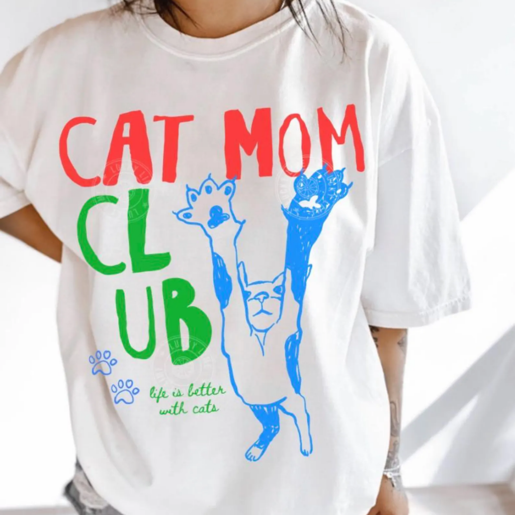 White t-shirt displaying bold text "CAT MOM CLUB" in red and green, features a playful blue cat illustration with raised paws.