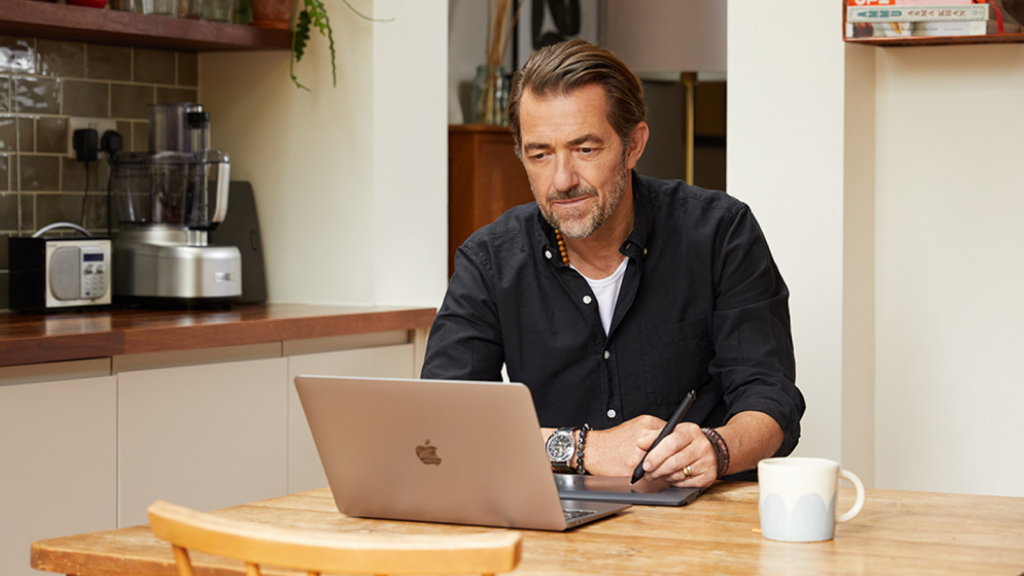 A man in a black shirt works intently on a laptop, creating custom flags, at a wooden kitchen table, stylus in hand.