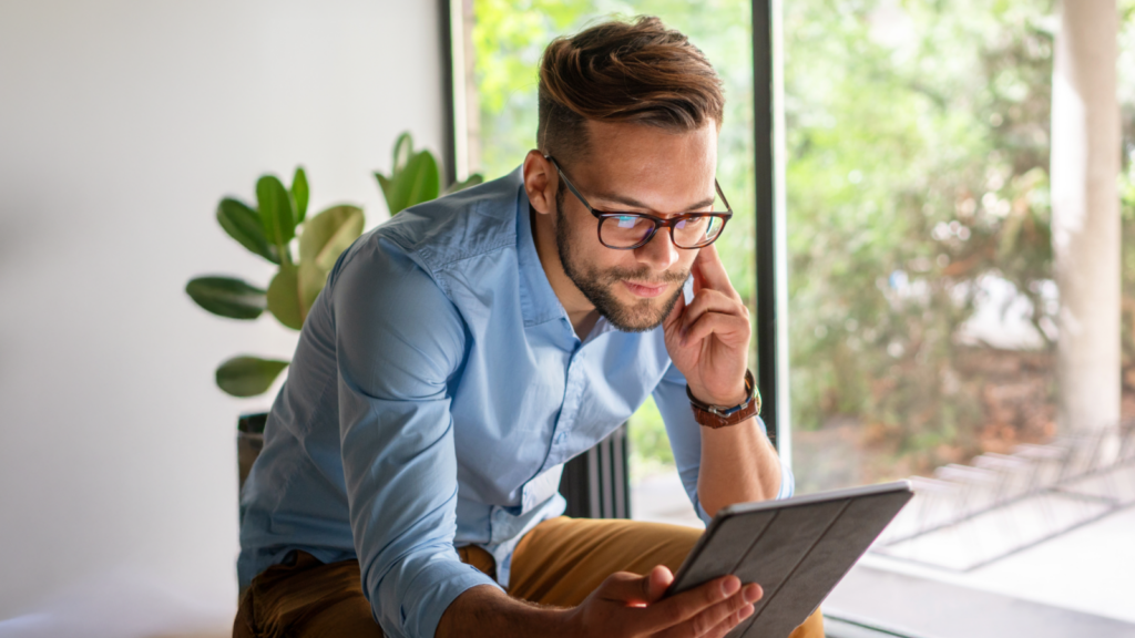 A man with glasses, wearing a blue shirt, sits by a window reading a tablet.