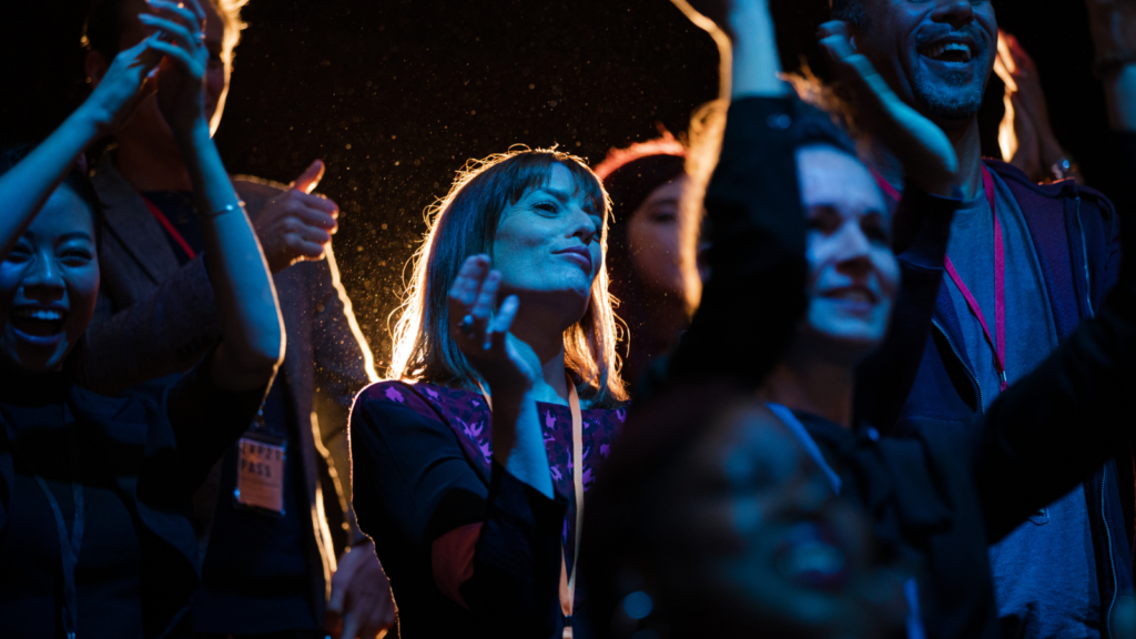 A joyous crowd celebrates at a concert, illuminated by blue and golden light. In focus, a woman gazes upwards.