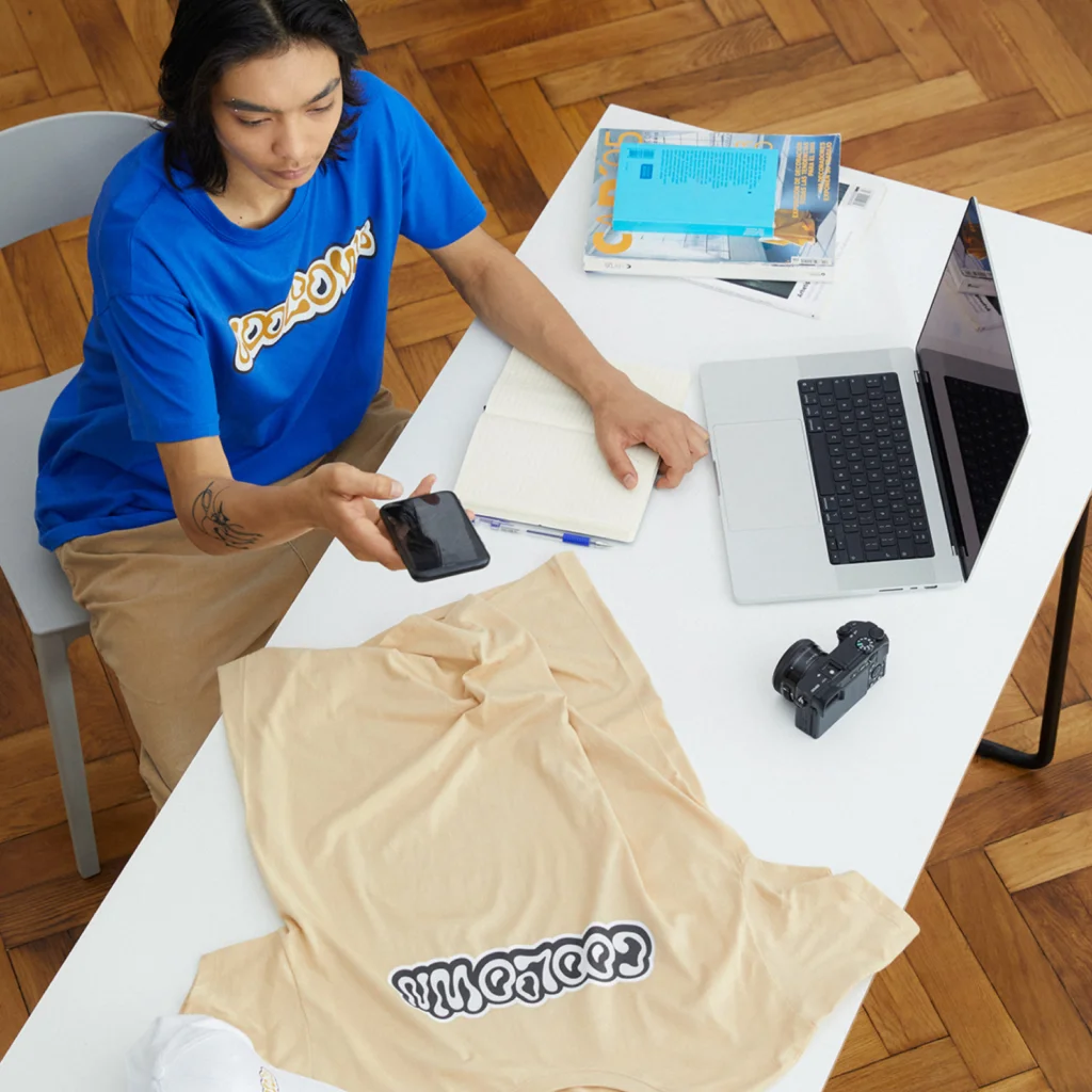 A man in a bright blue shirt and beige pants is seated at a white table, taking a picture of a beige t-shirts with a text print.